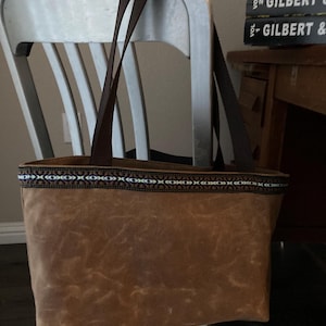 May include: A brown tote bag with dark brown straps. The bag has a decorative band with a geometric pattern in blue, white, and brown along the top edge. The bag is sitting on a silver chair.