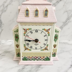 Könnte beinhalten: Eine Porzellan-Tischuhr in Hausform mit einem blassrosa Dach und drei Dachgauben. Das Zifferblatt zeigt schwarze römische Ziffern, den Markennamen Lenox und einen roten Sekundenzeiger. Blumenmuster verzieren die weiße Vorderseite und die Seiten.