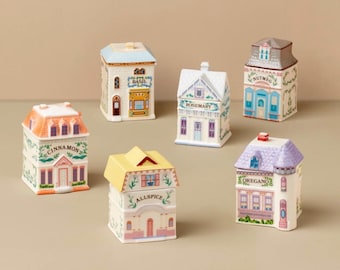 May include: A set of six ceramic spice jars shaped like miniature houses, each labeled with a specific spice name including Cinnamon, Basil, Rosemary, Nutmeg, Allspice, and Oregano. Each jar features a unique roof color and architectural details.