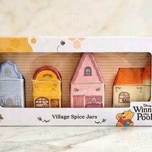 Könnte beinhalten: Ein Set aus vier hausförmigen Gewürzdosen aus Keramik in einer rechteckigen Schachtel. Die Dosen sind blau, gelb, rosa und cremefarben mit Winnie Puuh-Charakterdetails. Die Verpackung zeigt ein Wabenmuster, den Text Village Spice Jars und das Disney Winnie the Pooh-Branding.
