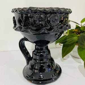 Puede incluir: Un cáliz de cerámica negra brillante con asa. El cáliz presenta un borde decorativo y diseños florales alrededor del cuenco. La base es redonda y el asa es curva. El cáliz mide aproximadamente 18 cm de altura.