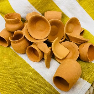 Puede incluir: Una colección de pequeñas piezas de cerámica de terracota sin esmaltar, que incluyen miniaturas de ollas, cuencos y fragmentos, dispuestas sobre un paño a rayas amarillas y blancas. La cerámica tiene un aspecto rústico y terroso.