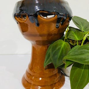 Puede incluir: Una maceta de cerámica hecha a mano con un diseño único. La maceta es de color marrón cálido con un esmalte marrón oscuro que gotea desde la parte superior. Tiene dos aberturas en forma de ojo y una base de pedestal. Una planta verde es visible en el fondo.