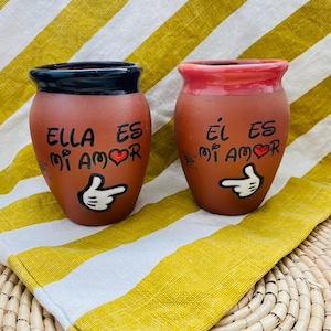 Puede incluir: Dos tazas de cerámica color terracota con bordes negros y rojos. Cada taza tiene un dedo que apunta dibujado a mano y las palabras "ELLA ES MI AMOR" y "ÉL ES MI AMOR" con un símbolo de corazón. Las tazas están sobre un paño a rayas amarillas y blancas.