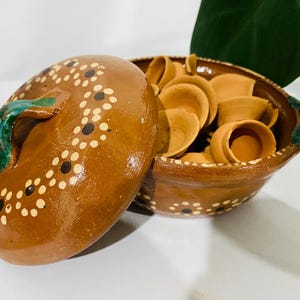 May include: A brown glazed clay pot with a matching lid, featuring hand-painted white and black dot patterns. The pot has green-glazed handles and is filled with several small, unglazed, light brown clay bowls or diya-style vessels.