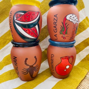 May include: Four terracotta ceramic vases with hand-painted designs. The vases feature images of a watermelon, a flamingo, a scorpion, and a pitcher. Each vase has a different coloured rim and base, and text in Spanish.