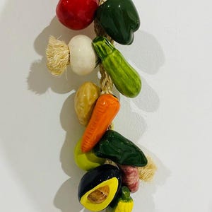 Puede incluir: Una guirnalda decorativa de cerámica con tomate rojo, pimiento verde, cebolla blanca, calabacín amarillo, zanahoria naranja, pimiento verde, aguacate y maíz amarillo. Las verduras están ensartadas en una cuerda natural.