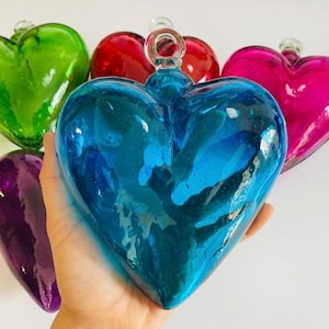 Puede incluir: Adornos de vidrio en forma de corazón en varios colores, incluyendo verde, rojo, rosa, azul y morado. Cada adorno tiene un lazo para colgar. Los adornos se muestran sobre un fondo blanco.