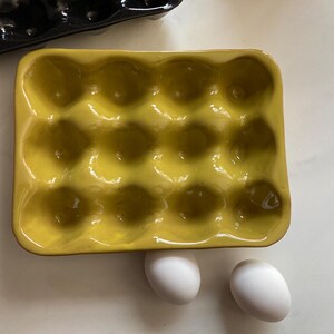 Pode incluir: Uma bandeja de ovos de cerâmica amarela com 12 reentrâncias, contendo dois ovos brancos. A bandeja é retangular com cantos arredondados e acabamento brilhante. Uma bandeja de ovos preta é parcialmente visível no fundo.