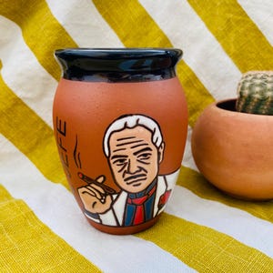 Puede incluir: Una taza de cerámica de color terracota con un borde negro. La taza presenta una ilustración pintada a mano de un hombre fumando un cigarro. La palabra "LIFE" está escrita en el lateral. Una pequeña maceta de terracota con un cactus está en el fondo.