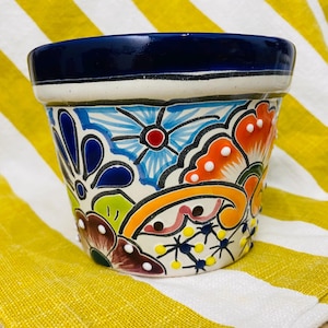 May include: A hand-painted ceramic flower pot with a vibrant floral design. The pot has a dark blue rim, white band, and colourful flowers in blue, orange, and green. The pot is set against a yellow and white striped background.