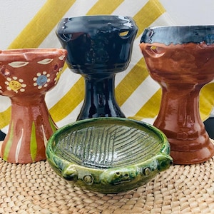 May include: Four handcrafted ceramic pieces: three pedestal cups and a small bowl. The cups are in shades of brown and black, with decorative details. The bowl is green with a textured surface. All pieces are on a woven mat.