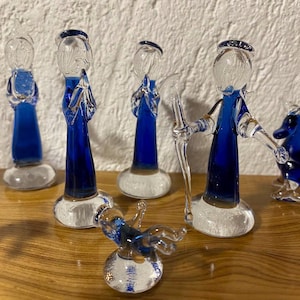 Op de afbeelding: Een verzameling glazen beeldjes die een kerststal uitbeelden. De figuren, waaronder herders, een engel en een os, zijn gemaakt van helder glas met blauwe accenten. De beeldjes staan op een houten oppervlak.
