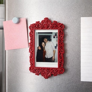 May include: A red, ornate picture frame with a photo of a couple, attached to a refrigerator with a magnet. A pink sticky note and a lined paper are also held by magnets. A small plant is visible.