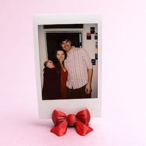 Puede incluir: Una foto Polaroid de dos personas, sostenida por un portafotos rojo en forma de lazo. El portafotos está hecho de un material brillante y se encuentra sobre una superficie rosa. La foto muestra a un hombre y una mujer sonriendo.