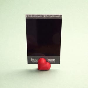 Könnte beinhalten: Eine schwarze Instax-Filmpackung mit dem Text "Don't put in mouth" und dem Instax-Logo, gehalten von einem kleinen, herzförmigen Objekt in Rot. Der Hintergrund ist hellgrün.