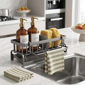 Puede incluir: Un organizador de fregadero de metal gris oscuro que contiene dos botellas de vidrio ámbar con dosificador etiquetadas Lemon Verbena Dish Soap y Hand Lotion, dos esponjas amarillas y un cepillo gris oscuro. Un paño a rayas verdes y blancas cuelga.