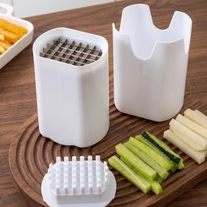 May include: A white french fry cutter with a stainless steel grid, a white container, and a white holder. Sliced cucumbers and potatoes are on a wooden cutting board. A tray of french fries is in the background.