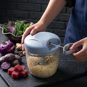 May include: A manual food chopper with a light blue lid and clear container, filled with chopped food. The chopper is being operated. Ingredients like purple potatoes, red beets, and herbs are arranged nearby.