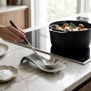 Può includere: Un poggia mestolo in ceramica grigio chiaro dal design curvo e scanalato che sostiene un mestolo in acciaio inossidabile con manico in legno marrone scuro. Il poggia mestolo è posizionato su un piano in marmo accanto a una pentola in ghisa.