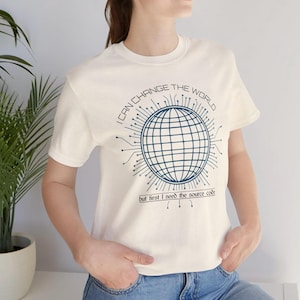 Peut inclure: T-shirt crème avec un motif de globe bleu entouré de lignes en forme de circuits. Le texte "I CAN CHANGE THE WORLD" est au-dessus du globe, avec "but first I need the source code" en dessous.