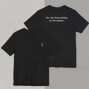 Op de afbeelding: Twee zwarte t-shirts met witte tekst. Eén shirt heeft een puntkomma op de voorkant, de andere de zin "The only thing holding my life together." op de achterkant. Beide zijn shirts met korte mouwen.
