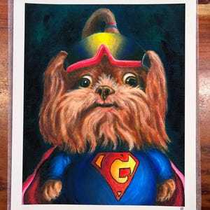 Peut inclure: Une impression d'une peinture représentant un chien brun à poils longs portant un costume de super-héros bleu avec un emblème rouge et jaune sur la poitrine, une cape rouge et un casque sombre avec des lunettes roses sur fond sombre.