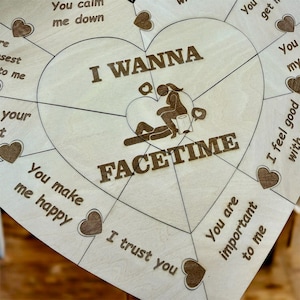 May include: A wooden heart-shaped game board with the words "I WANNA FACETIME" in a bold, brown font. The board is divided into sections with phrases like "You make me happy" and "I trust you". Small heart shapes are scattered throughout.