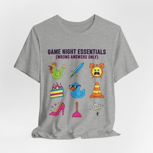 Può includere: Una maglietta grigia con la scritta "GAME NIGHT ESSENTIALS (WRONG ANSWERS ONLY)". La maglietta presenta illustrazioni di cartoni animati colorati di un pollo, una spada, una sveglia, una torta, un'anatra, un cono stradale, un tacco alto, uno sturalavandini e un UFO.