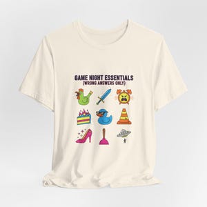 Può includere: T-shirt color crema con la scritta "GAME NIGHT ESSENTIALS (WRONG ANSWERS ONLY)" e illustrazioni di cartoni animati colorati. Le illustrazioni includono un pollo, una spada, una sveglia, una torta, un'anatra, un cono stradale, un tacco alto, uno sturalavandini e un UFO.