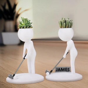 May include: Two white ceramic golf-themed planters. Each planter features a figure with a spherical head holding a golf club. One holds a succulent, the other a cactus. The name "JAMES" is on a small sign on one planter.