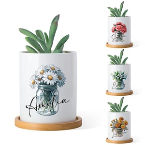 May include: White ceramic planter with a succulent plant. The planter features a watercolor illustration of daisies in a mason jar with the name "Amelia" in script. Includes a small, round, wooden saucer.