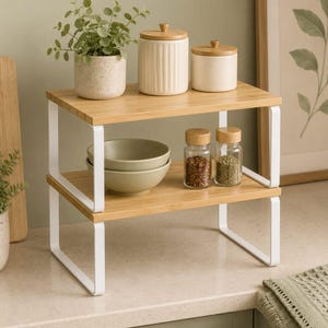 May include: A two-tiered bamboo and white metal shelf unit. The top shelf holds a potted plant, two ceramic jars with wooden lids. The bottom shelf has two bowls and two spice jars. The unit is 12 inches wide.