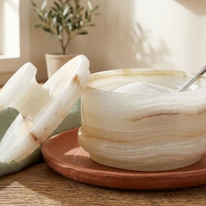 Pode incluir: Uma açucareiro decorativo com tampa, feito de ônix branco e bege. A tigela está sobre um prato cor de terracota, e uma pequena colher de prata está dentro. Um guardanapo de linho verde e canecas verdes estão no fundo.