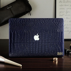 May include: A dark blue laptop cover with a textured crocodile skin pattern, featuring a central white Apple logo and the gold-colored initials BH printed in the bottom right corner. The cover is designed to fit a laptop computer.