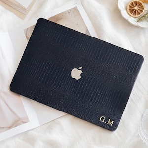 Puede incluir: Una funda para portátil de color azul marino oscuro con un patrón texturizado de piel de caimán. La funda presenta un recorte central para el logotipo plateado de Apple y las iniciales doradas G.M impresas en la esquina inferior derecha.