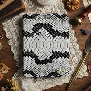 May include: A black and white snakeskin-patterned journal with gold initials. The journal is on a wooden surface with a vintage pocket watch, a feather quill, and other decorative items. The journal's cover has a reptile scale design.