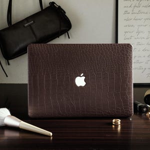 May include: A laptop computer featuring a dark brown, textured crocodile-patterned skin on its lid. The iconic white Apple logo is illuminated in the center of the cover. The device is positioned on a dark surface alongside a makeup brush.