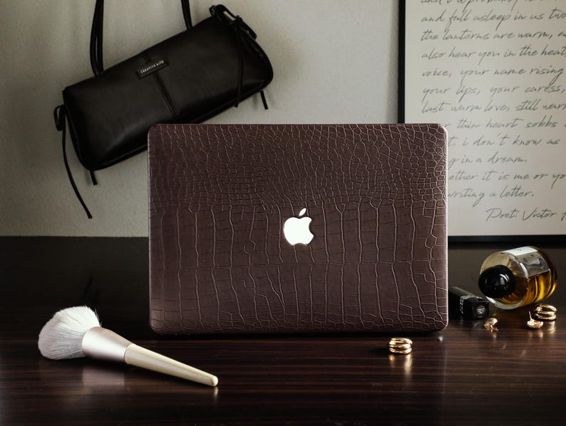 May include: A laptop computer featuring a dark brown, textured crocodile-patterned skin on its lid. The iconic white Apple logo is illuminated in the center of the cover. The device is positioned on a dark surface alongside a makeup brush.