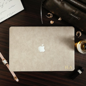 Peut inclure: Un autocollant pour ordinateur portable beige avec un motif texturé imitation alligator appliqué sur le couvercle d'un ordinateur argenté. L'autocollant présente les initiales dorées H.A. imprimées dans le coin inférieur droit, centré autour du logo Apple.
