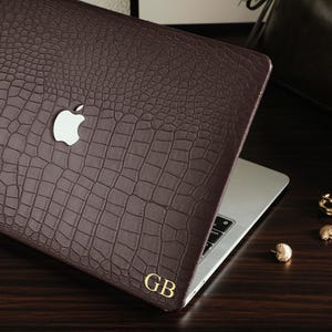 May include: A dark brown laptop cover with a textured crocodile skin pattern, featuring a cutout for the silver Apple logo and the gold-colored initials GB printed in the bottom right corner, applied to a silver laptop.