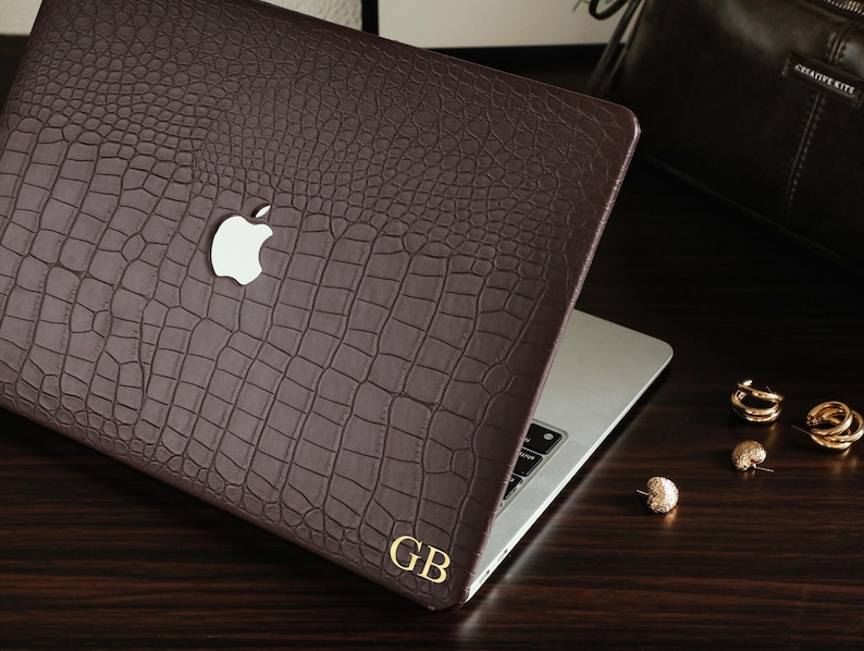May include: A dark brown laptop cover with a textured crocodile skin pattern, featuring a cutout for the silver Apple logo and the gold-colored initials GB printed in the bottom right corner, applied to a silver laptop.