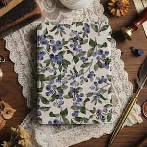 May include: A notebook featuring a watercolour design of blueberries and green leaves on a cream background. The notebook has gold initials in the corner. It rests on a wooden surface with a lace doily, a pocket watch, and a quill pen.