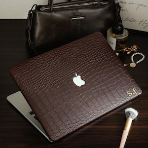 May include: A dark brown laptop cover with a textured crocodile skin pattern and a cutout for the Apple logo. The cover features the gold embossed initials S.F. in the bottom right corner, providing a sophisticated and personalized appearance.