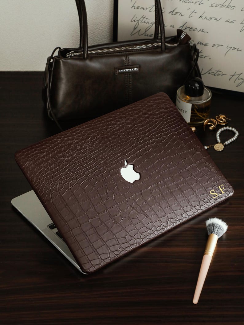 May include: A dark brown laptop cover with a textured crocodile skin pattern and a cutout for the Apple logo. The cover features the gold embossed initials S.F. in the bottom right corner, providing a sophisticated and personalized appearance.