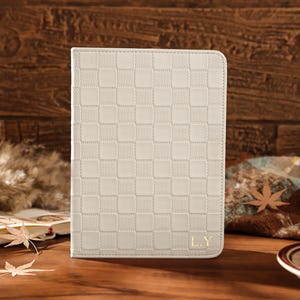 May include: A cream-colored tablet case with a checkered pattern and the initials "L.Y." in gold. The case is on a wooden surface with autumn leaves and a cup of coffee in the background. The case is designed to protect a tablet.