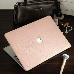 May include: A light pink laptop cover with a textured crocodile skin pattern and a cutout for the Apple logo. The cover features the gold-colored initials S.A. printed in the bottom right corner, providing a customized look for the device.