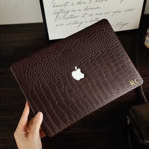 May include: A dark brown laptop cover with a textured crocodile skin pattern. The cover features a central cutout for the Apple logo and the gold-colored initials RC printed in the bottom right corner, providing a protective layer for the device.