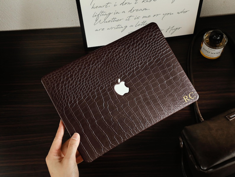 May include: A dark brown laptop cover with a textured crocodile skin pattern. The cover features a central cutout for the Apple logo and the gold-colored initials RC printed in the bottom right corner, providing a protective layer for the device.