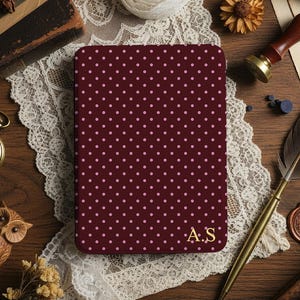 May include: A burgundy notebook with a pink polka dot pattern and gold initials "A.S" is displayed on a wooden surface. The scene includes a pocket watch, quill, wax seal, and lace.